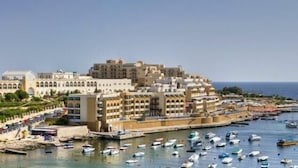 A coastal town with a prominent building complex, marina with numerous boats, and a clear view of the sea. - Verdi St Georges Bay Marina (St. Julian's)