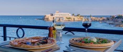 A table with two pizzas and glasses of wine, overlooking a body of water with a building in the background.