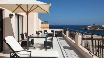 Un balcón con mobiliario exterior, una mesa con un florero y vistas al mar y a un edificio.
