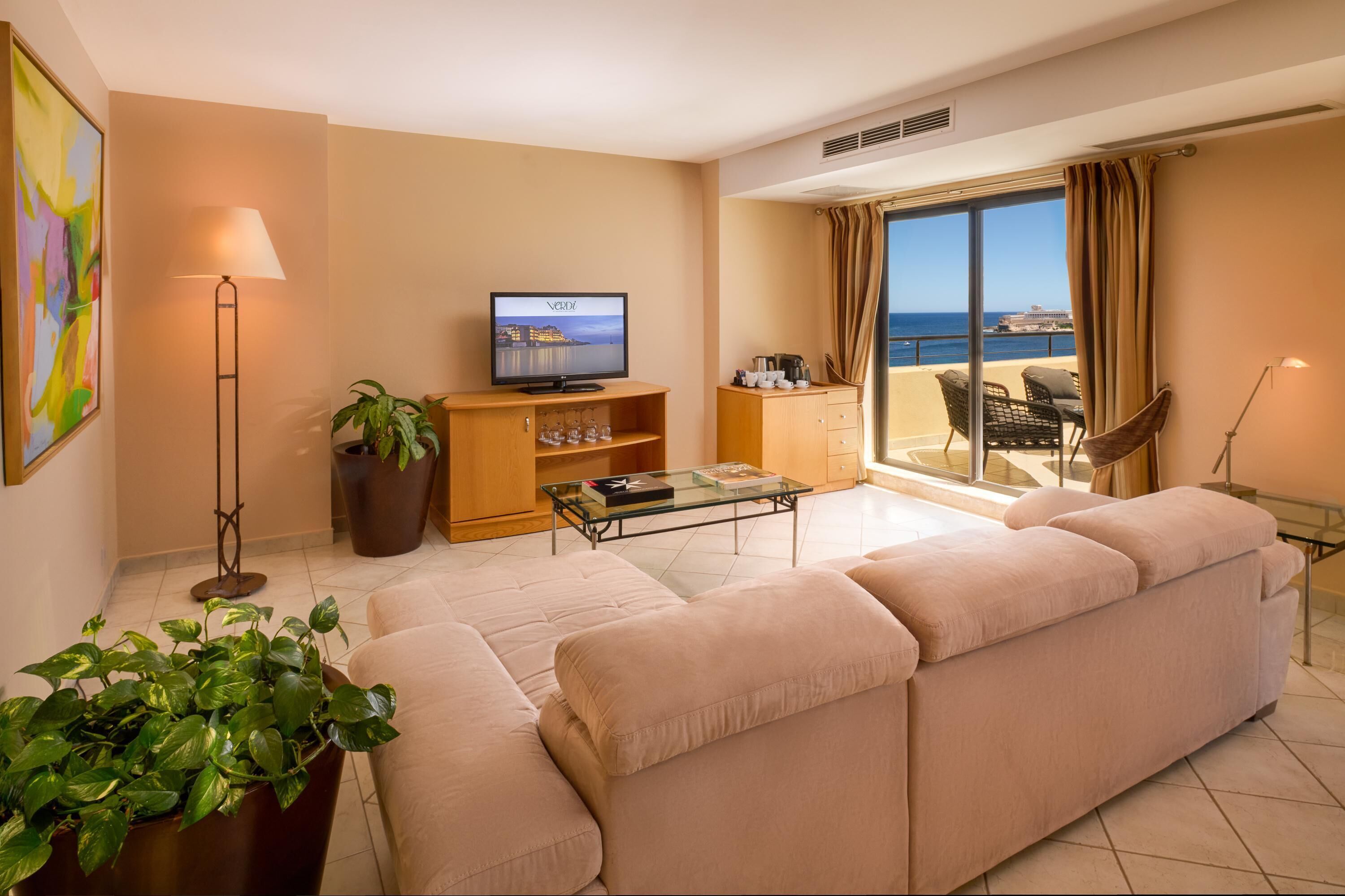 A living room with a sofa, a coffee table, a TV, a plant, a bookshelf, and a balcony with a view of the sea.