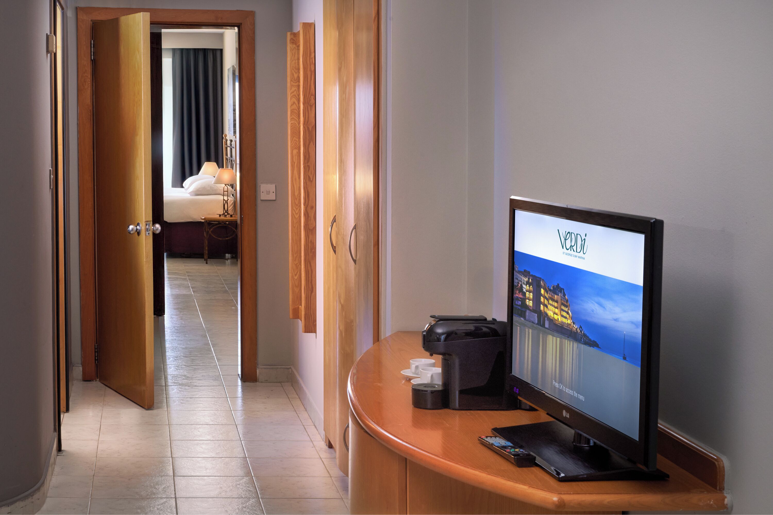 A hotel room with a wooden desk, a television displaying a cityscape, and a bed with a nightstand.