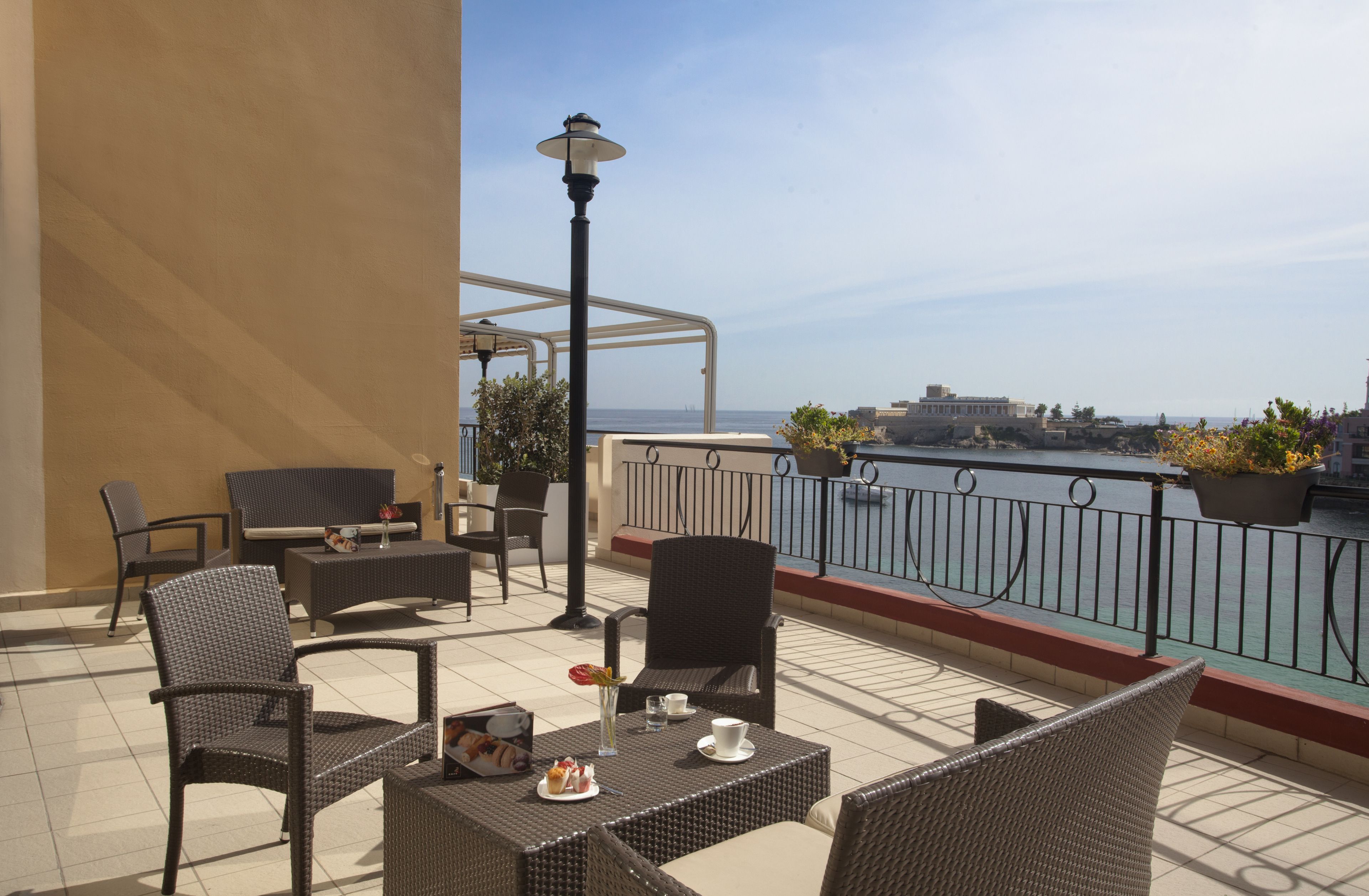 Una terraza con mobiliario de mimbre, una mesa con un centro de mesa floral y vistas al mar y al paisaje urbano.