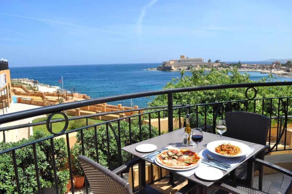 Un balcon avec une table dressée pour deux personnes, offrant une vue sur la côte et un bâtiment visible au loin.