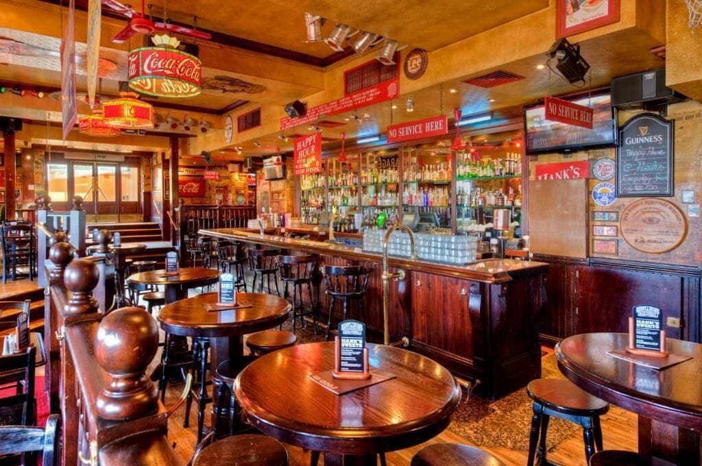 A bar with wooden tables and chairs, a counter with various bottles, and signs for Coca-Cola and Guinness.