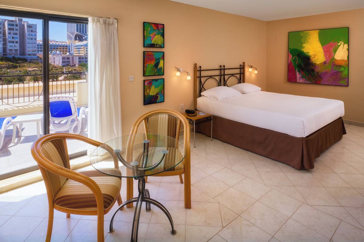 A hotel room with a bed, a glass table with chairs, a balcony with a view of buildings, and three abstract paintings on the wall.
