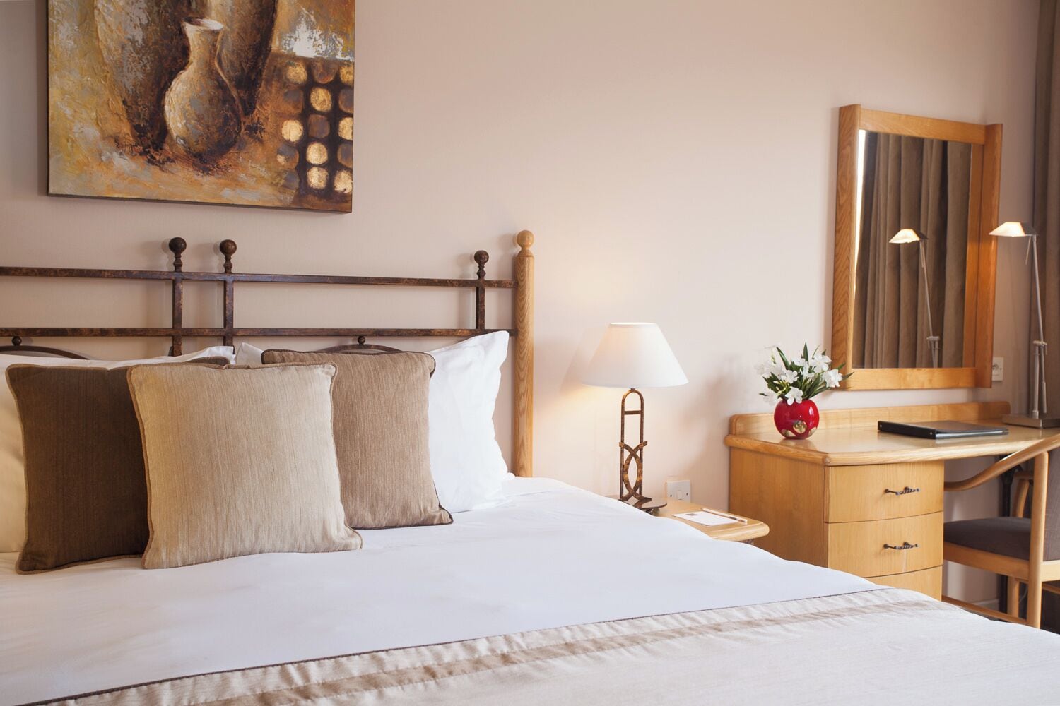 A neatly made bed with two pillows, a bedside table with a lamp, a small red vase with flowers, and a wooden desk with a mirror.