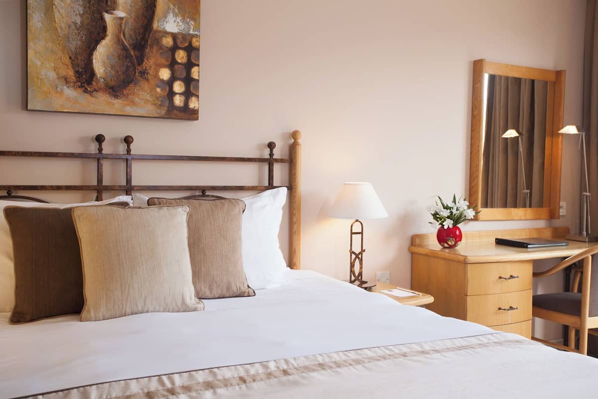 A neatly made bed with two pillows, a bedside table with a lamp, a small red vase with flowers, and a wooden desk with a mirror.