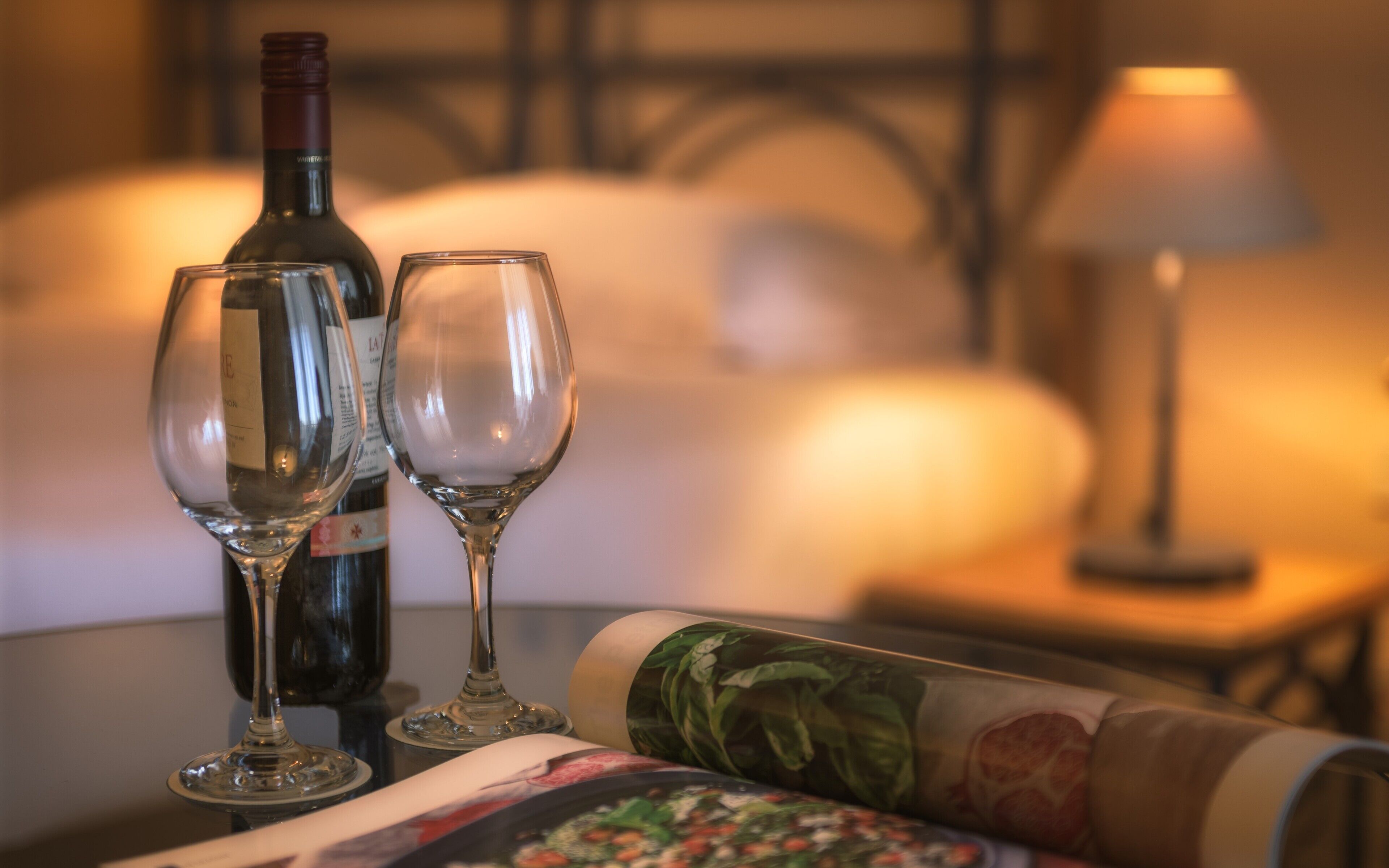 Une bouteille de vin et deux verres sur une table, avec une lampe et un lit en arrière-plan.