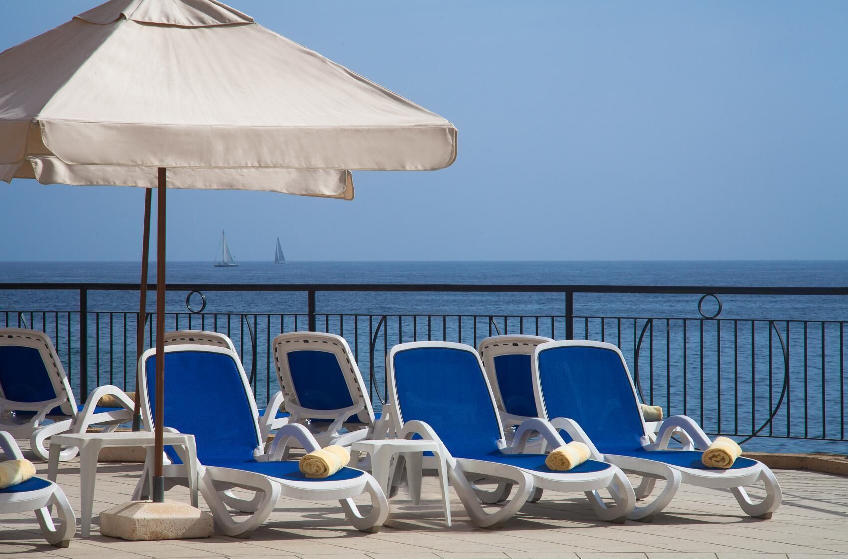 Une rangée de chaises longues blanches, dotées de coussins bleus et surplombées d’un parasol beige, se trouve sur une terrasse offrant une vue sur la mer et les voiliers.
