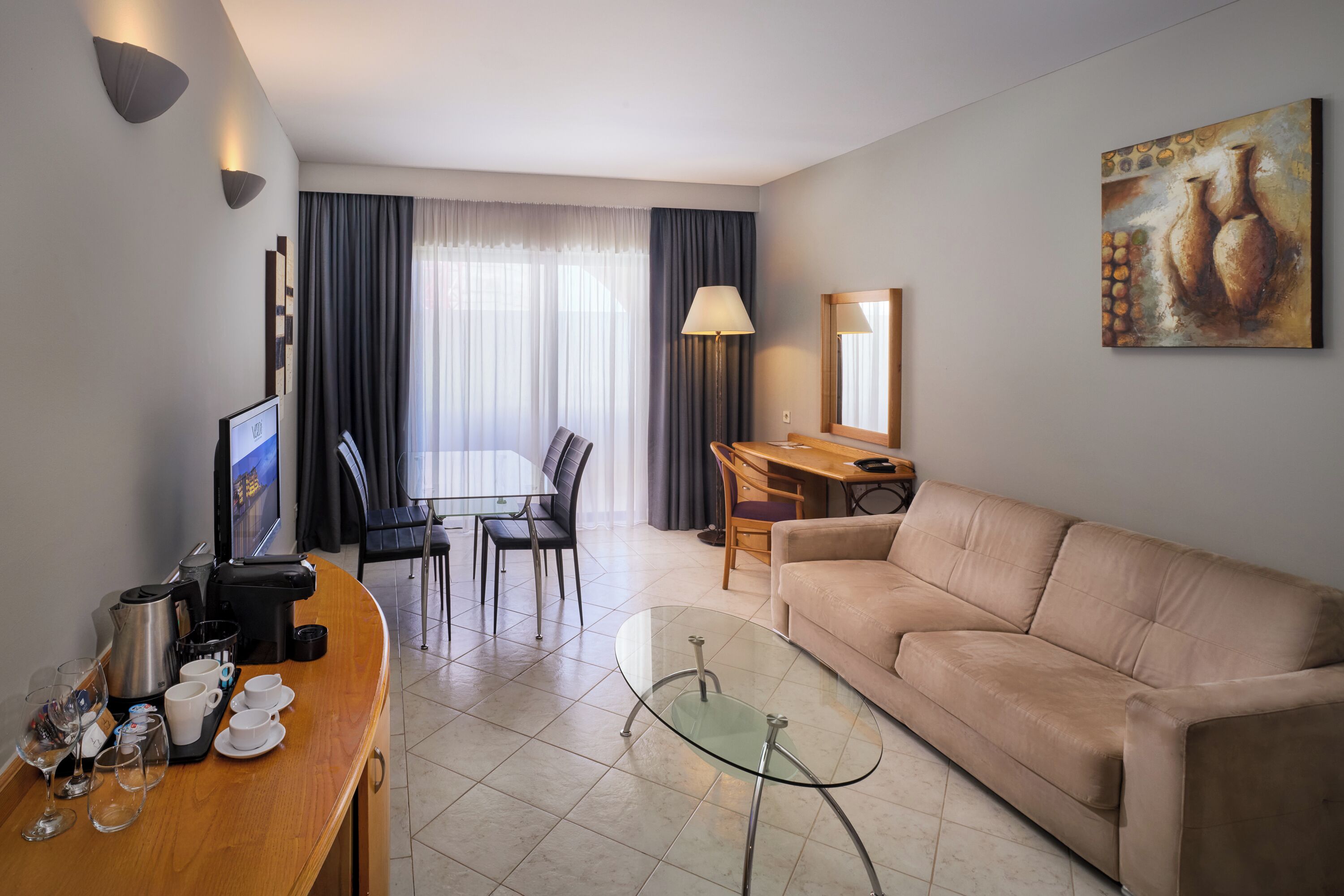 Una habitación de hotel moderna con sofá, una mesa de centro de vidrio, un área de comedor con mesa y sillas, un televisor y un cuadro en la pared.