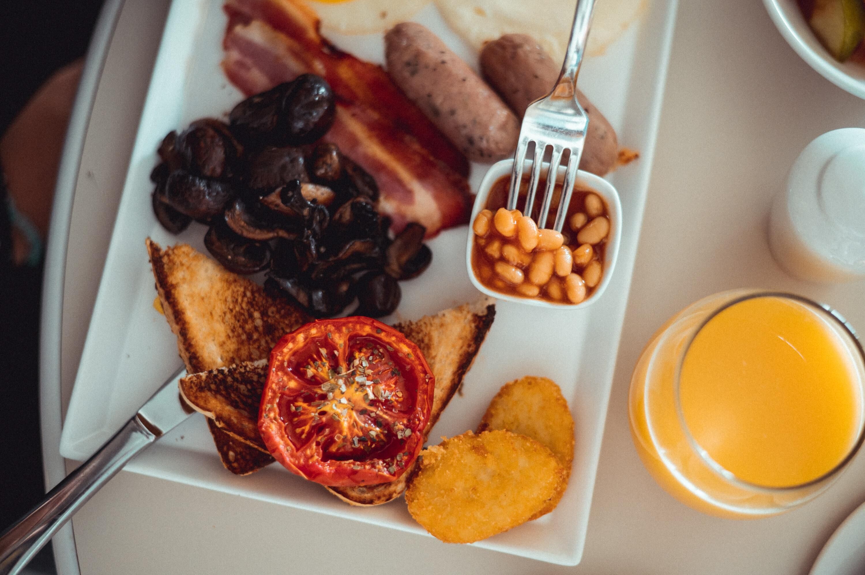 A breakfast plate with toast, mushrooms, bacon, beans, a tomato slice, hash browns, and a glass of orange juice.
