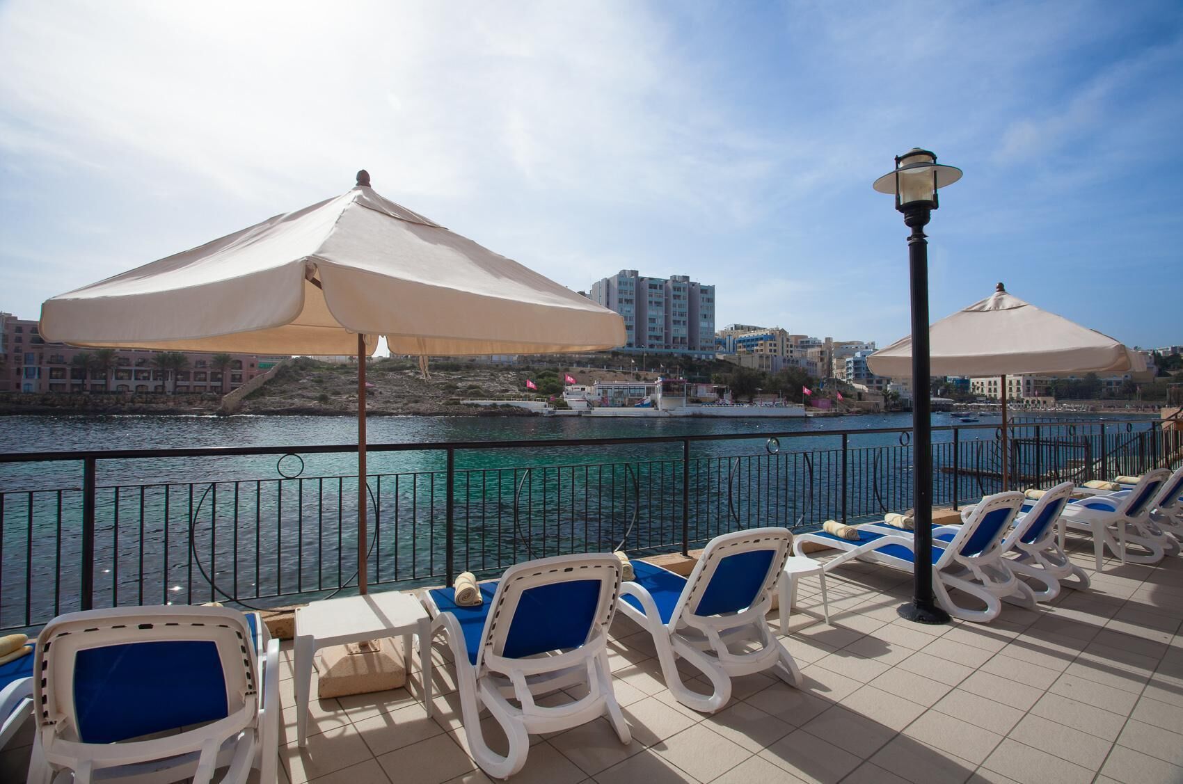A waterfront terrace with lounge chairs and umbrellas, overlooking a body of water and buildings in the background.