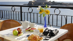 A breakfast table with croissants, fruits, and a camera, overlooking a coastal view with a historic building. - Verdi St Georges Bay Marina (St. Julian's)