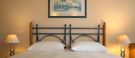 A bed with white linens, a blue metal headboard, two bedside lamps, and a framed painting above the bed.