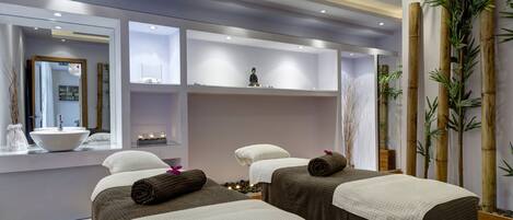 A modern spa room with two beds, a large mirror, a sink, and shelves with decorative items.
