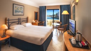 A hotel room with a large bed, a desk with a computer, a TV, and a view of the sea. - Verdi St Georges Bay Marina (St. Julian's)