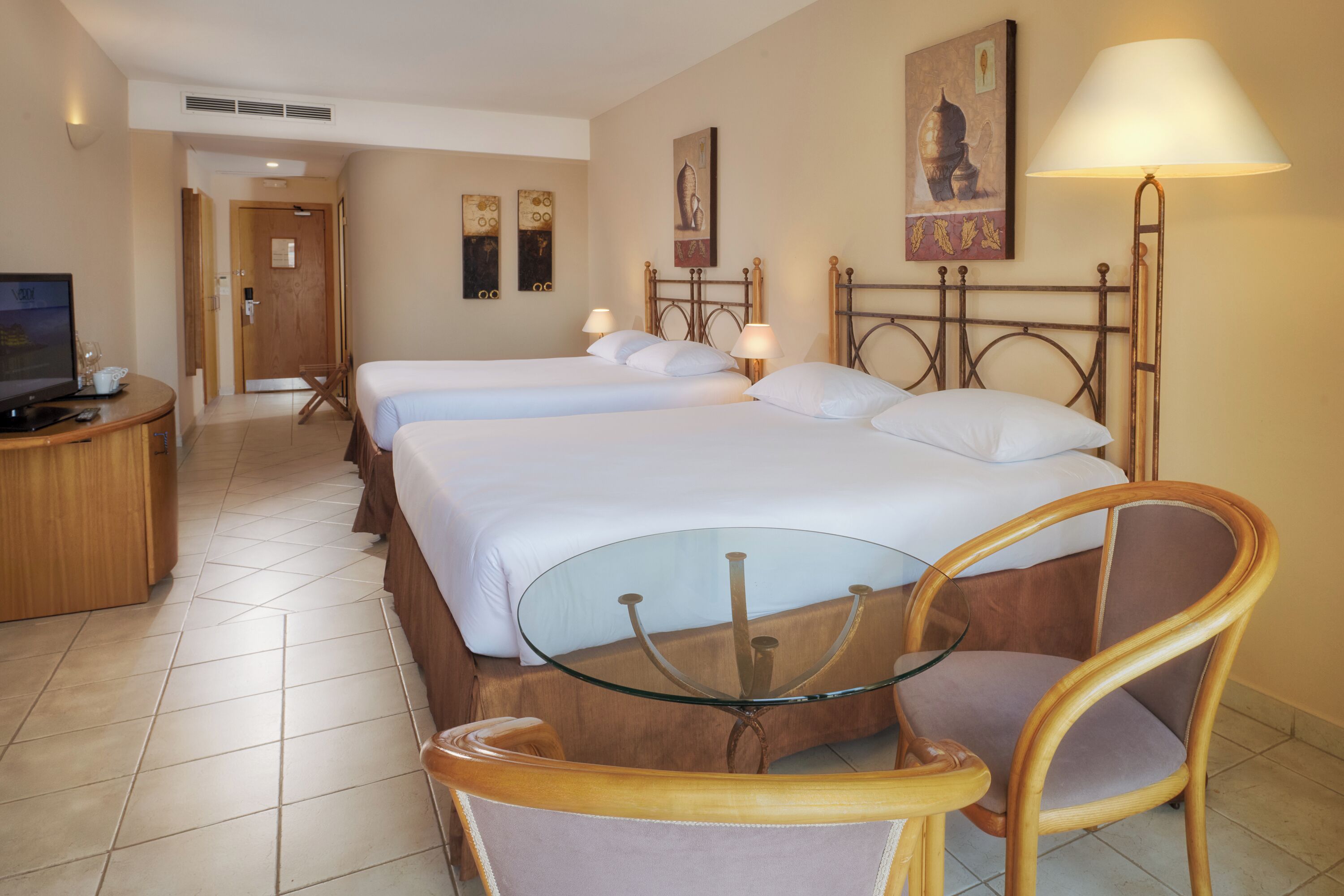 Habitación de hotel con dos camas, una mesa de vidrio, una silla de madera y un televisor sobre un mueble de madera.