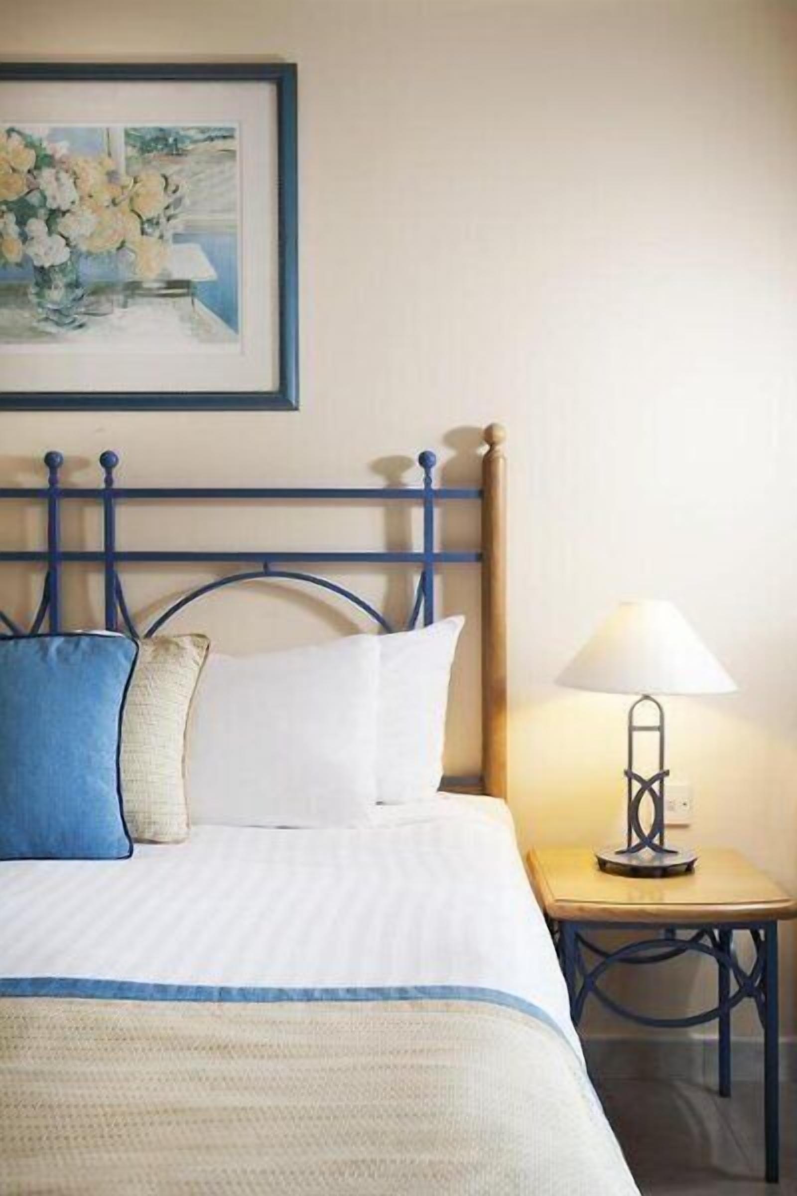 Una cama bien hecha con un cabecero azul, una mesita de noche con una lámpara y un cuadro enmarcado en la pared.