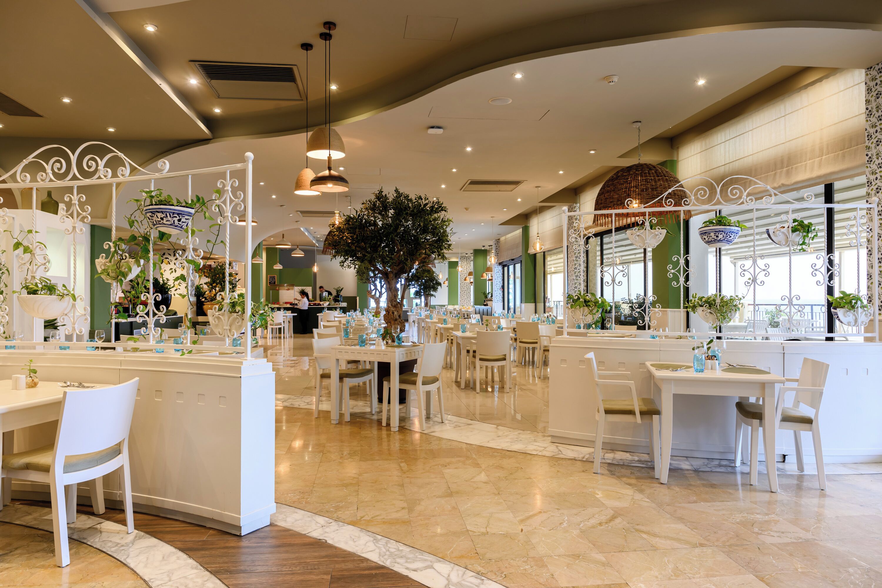 Een ruim restaurant met witte meubels en een centrale boom, met een modern design, gebogen plafonds en decoratieve verlichting.