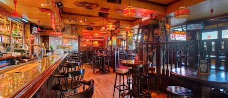 A bar with wooden floors and multiple stools, a counter with bottles, and various signs.