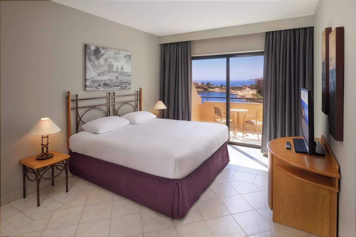 A hotel room with a bed, a desk with a computer, a TV, a painting, and a balcony with a view of the sea.