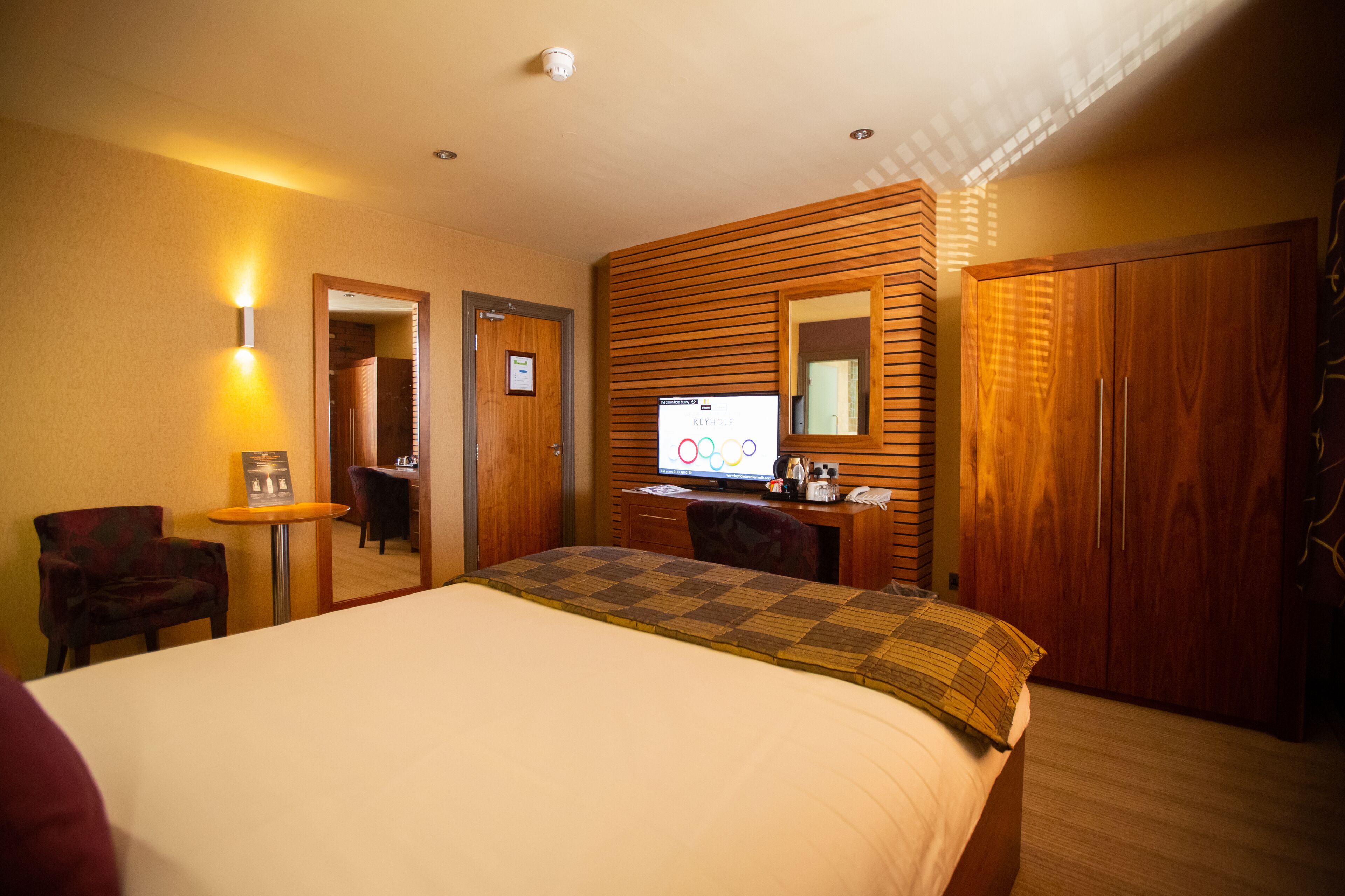 A hotel room with a large bed, a desk with a computer, a chair, a small table with a lamp, and a wardrobe.