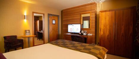A hotel room with a large bed, a desk with a computer, a chair, a small table with a lamp, and a wardrobe.