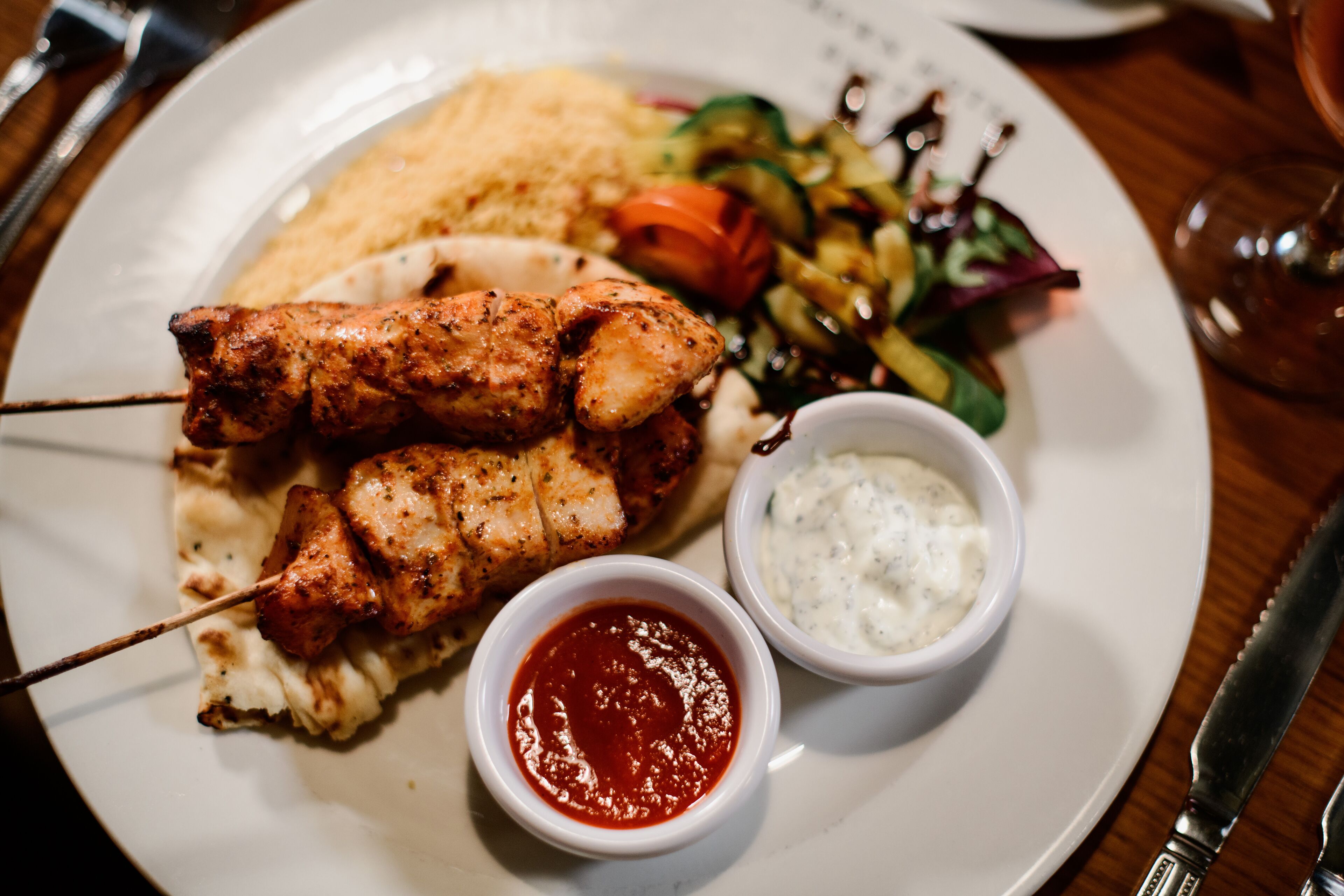 Un plato con brochetas de pollo a la parrilla, ensalada, pan plano y dos salsas para mojar.