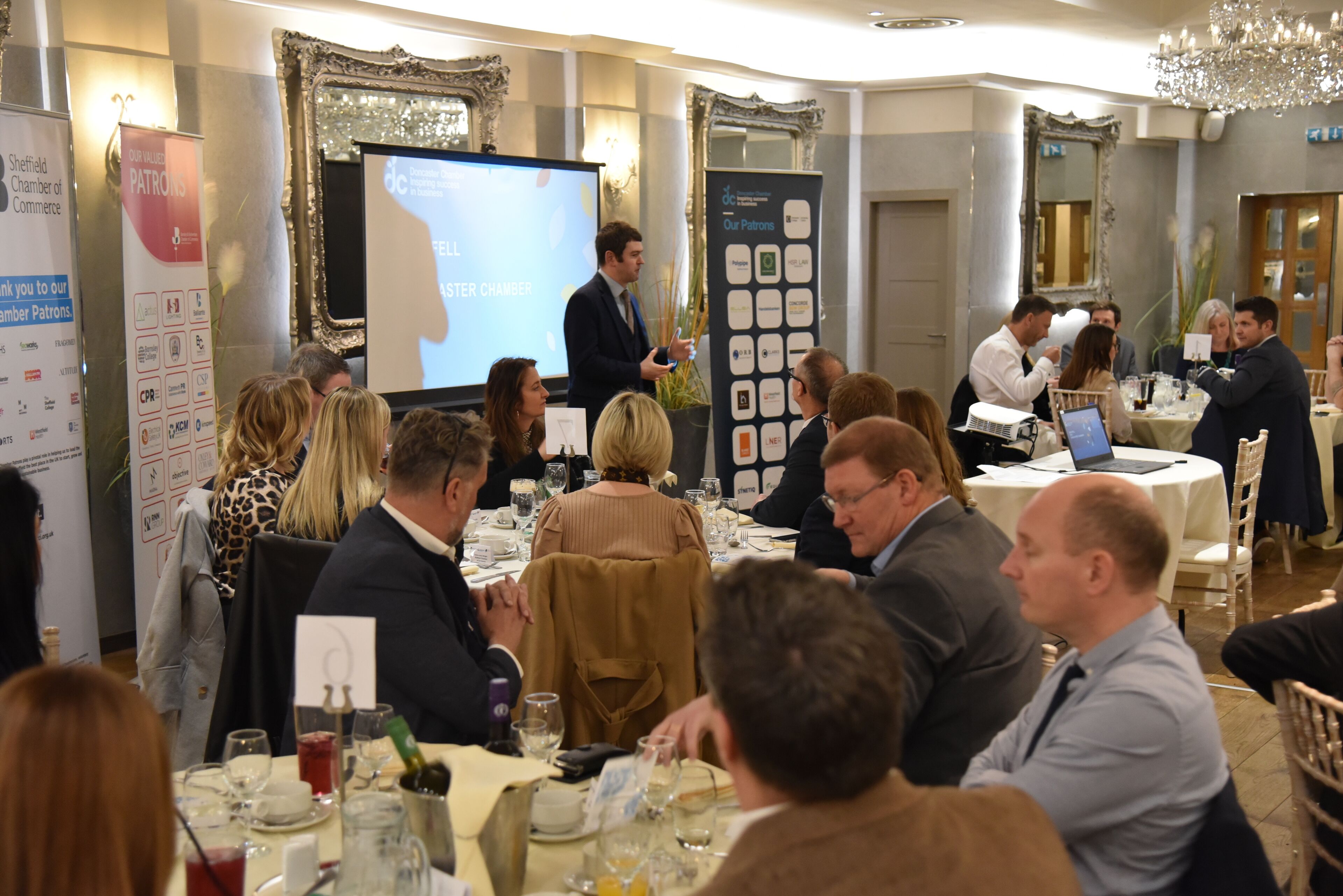 A formal event with attendees seated at round tables, a speaker presenting at the front, and a banner displaying 'Patrons' and 'Sheffield Chamber of Commerce'.