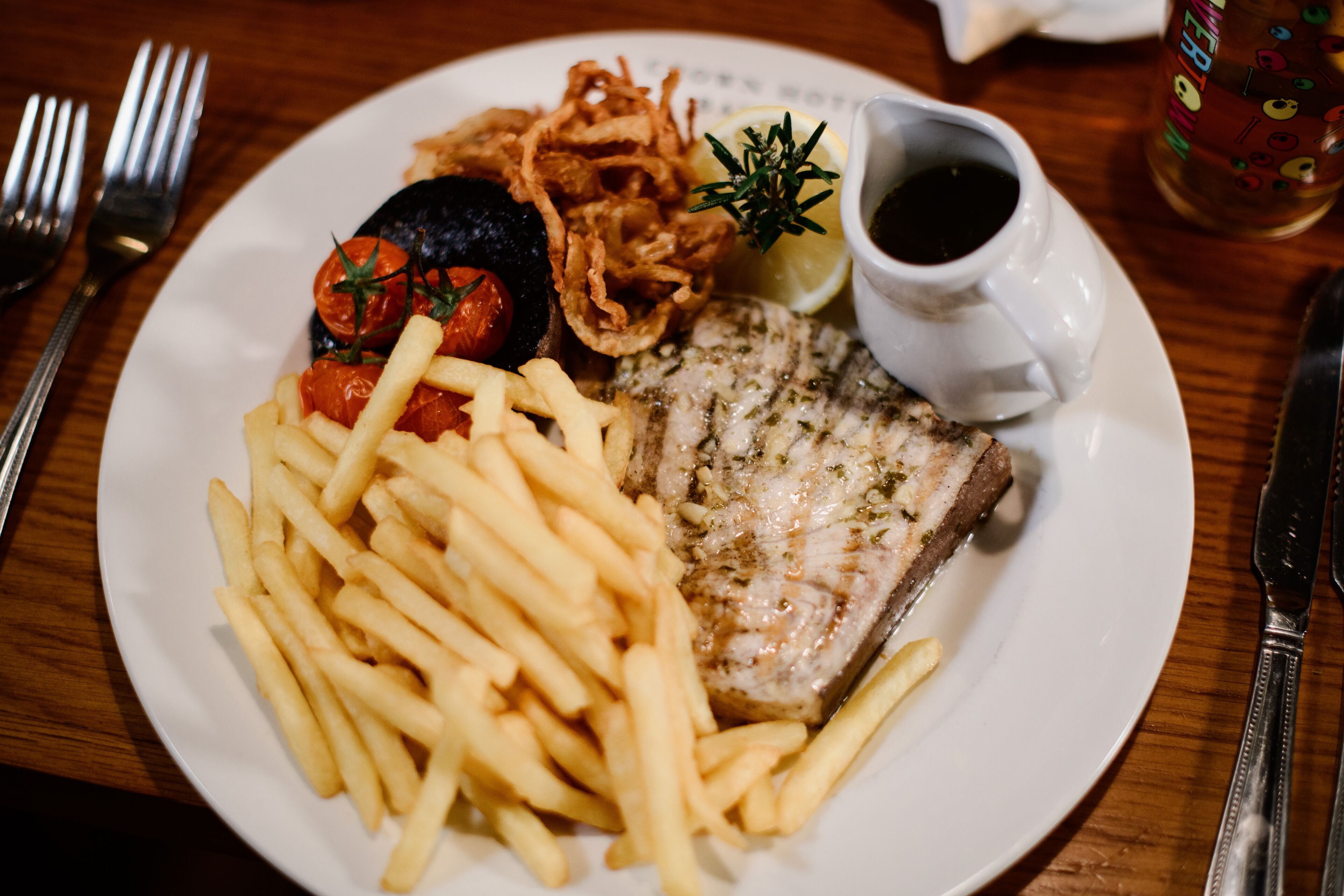 Un plato con un pescado a la parrilla, papas fritas, tomates cherry y aros de cebolla, acompañado de una pequeña taza de salsa y una rodaja de limón.