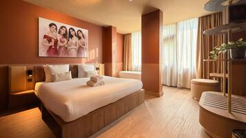 A modern hotel room with a large bed, a wall-mounted painting, a bedside table, a chair, and a plant.