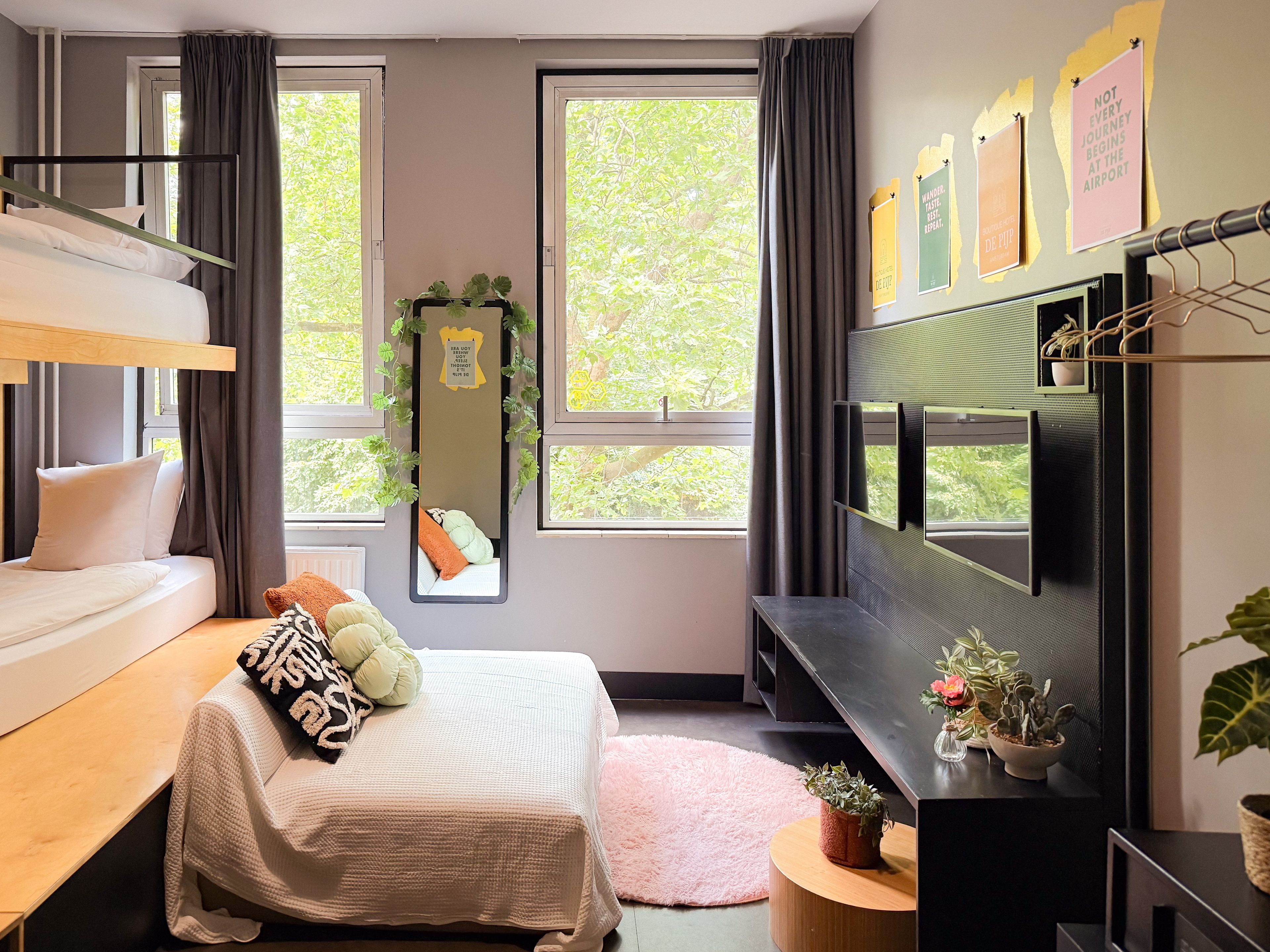 A modern hotel room with a bunk bed, a flat-screen TV, a small desk, and a large window offering a view of trees.