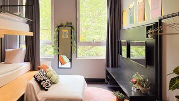 A modern hotel room with a bunk bed, a flat-screen TV, a small desk, and a large window offering a view of trees.