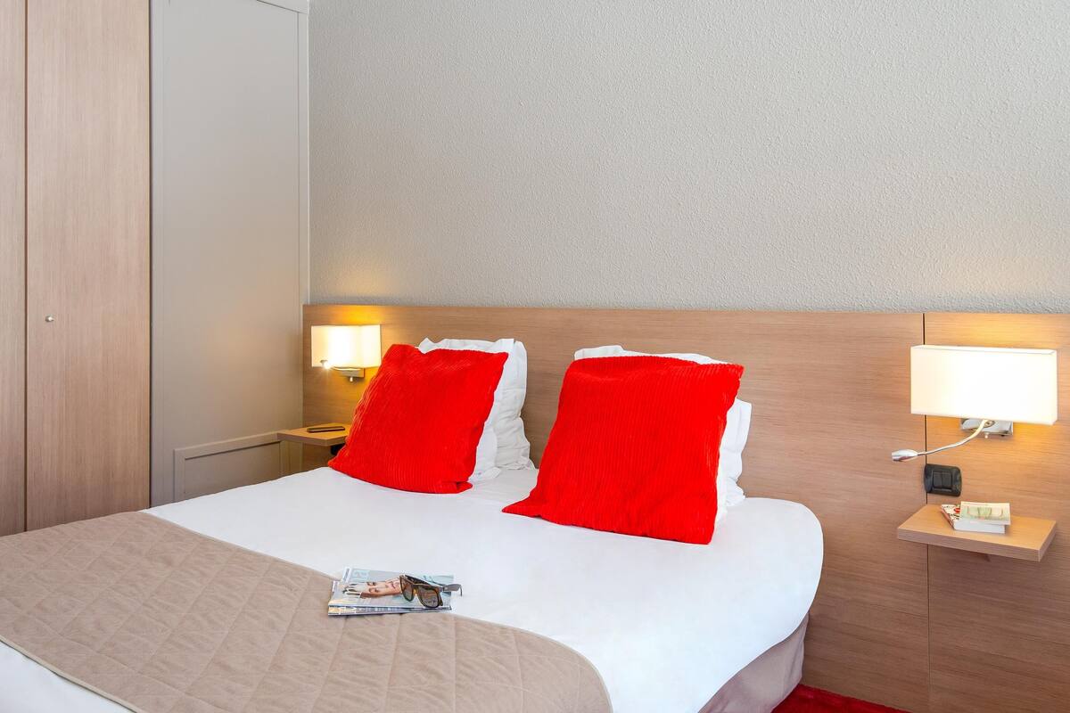 A hotel room with a bed, two red pillows, a bedside table with a lamp, and a magazine on the bed.