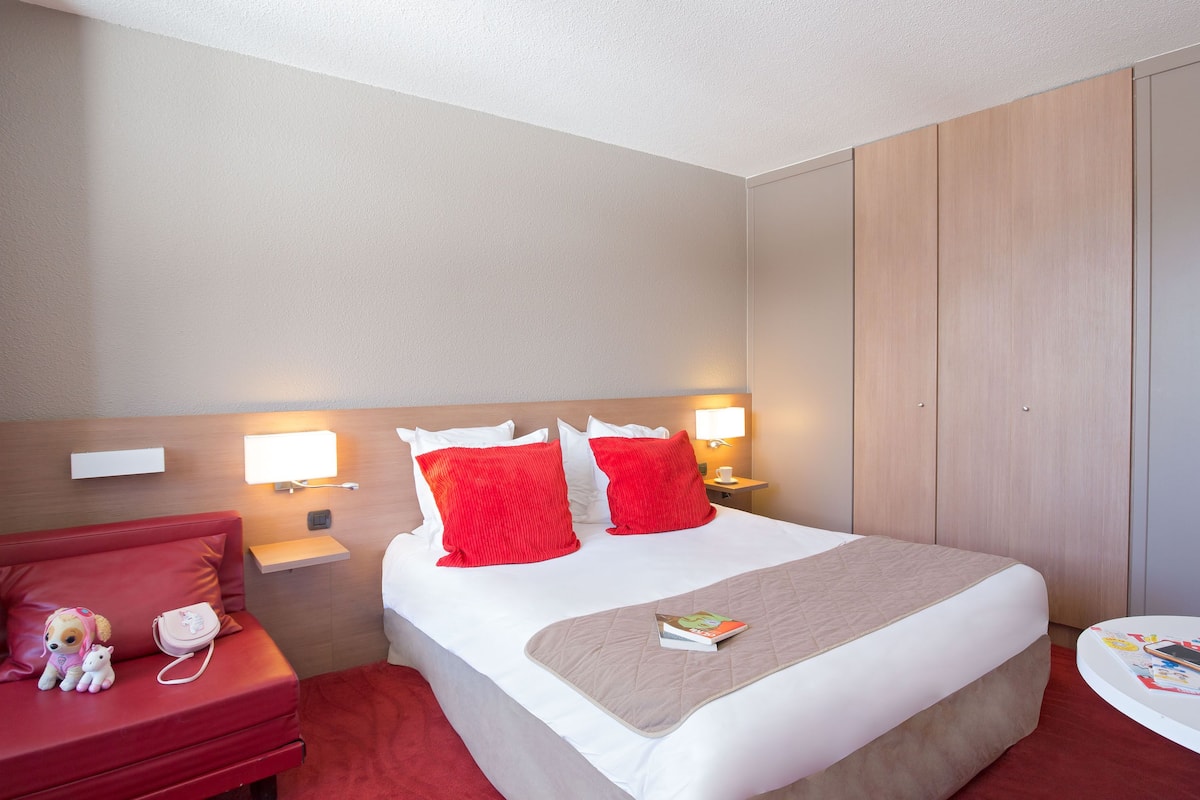 A hotel room with a bed, a red sofa, a small table, and a nightstand with a lamp.