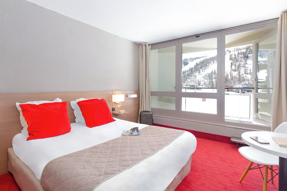 Habitación de hotel con una cama grande, almohadas rojas y blancas, una colcha acolchada, una mesita de noche con lámpara y vistas a un paisaje nevado a través de un ventanal amplio.