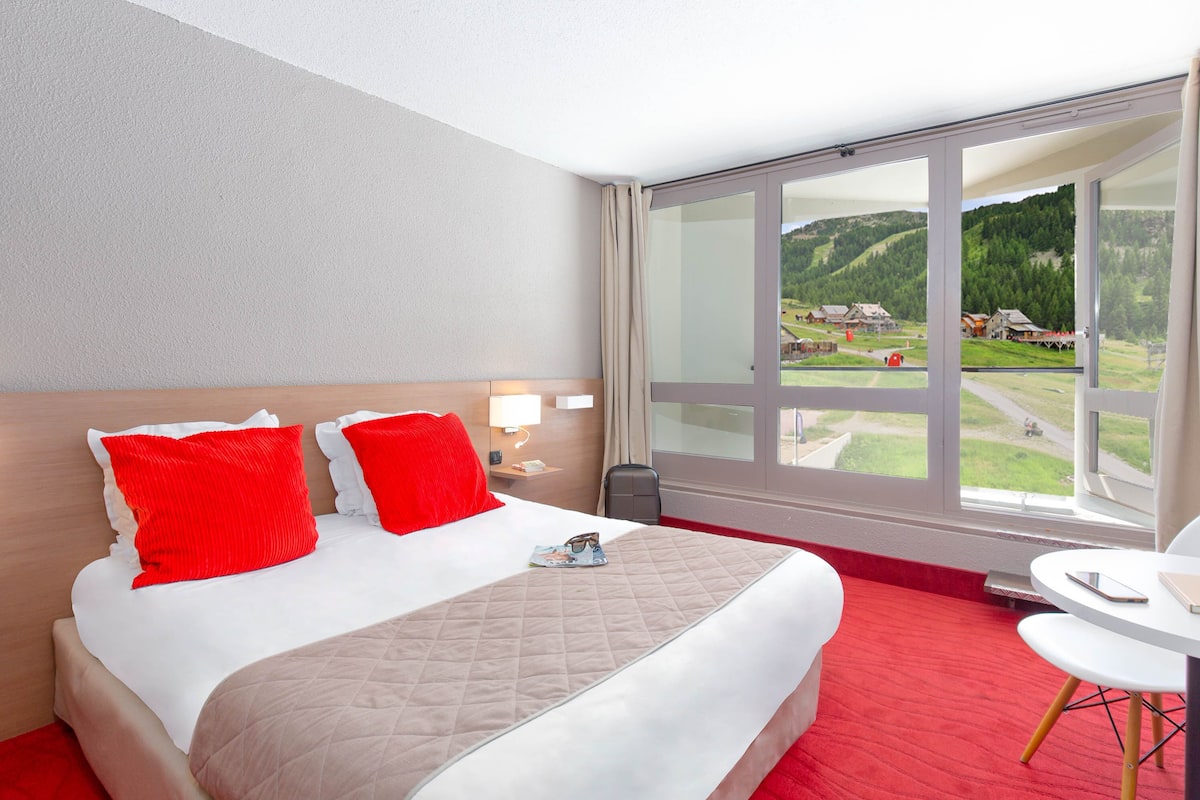 A hotel room with a large bed, red and white pillows, a bedside table with a lamp, and a view of a green landscape through a large window.
