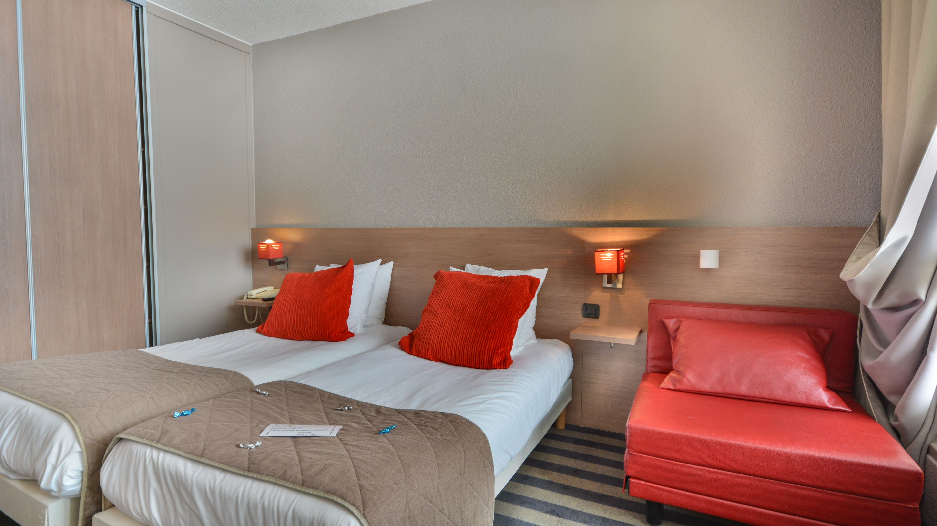 Camera d'albergo con un letto matrimoniale, due cuscini rossi, un divano rosso e un comodino con una lampada.