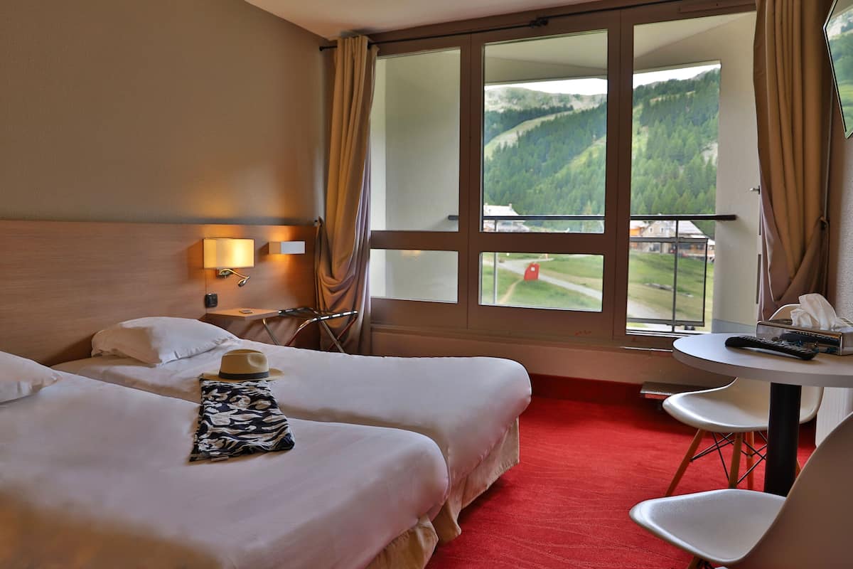A hotel room with two beds, a small table with a chair, a window with a view of a green landscape, and a lamp on the wall.