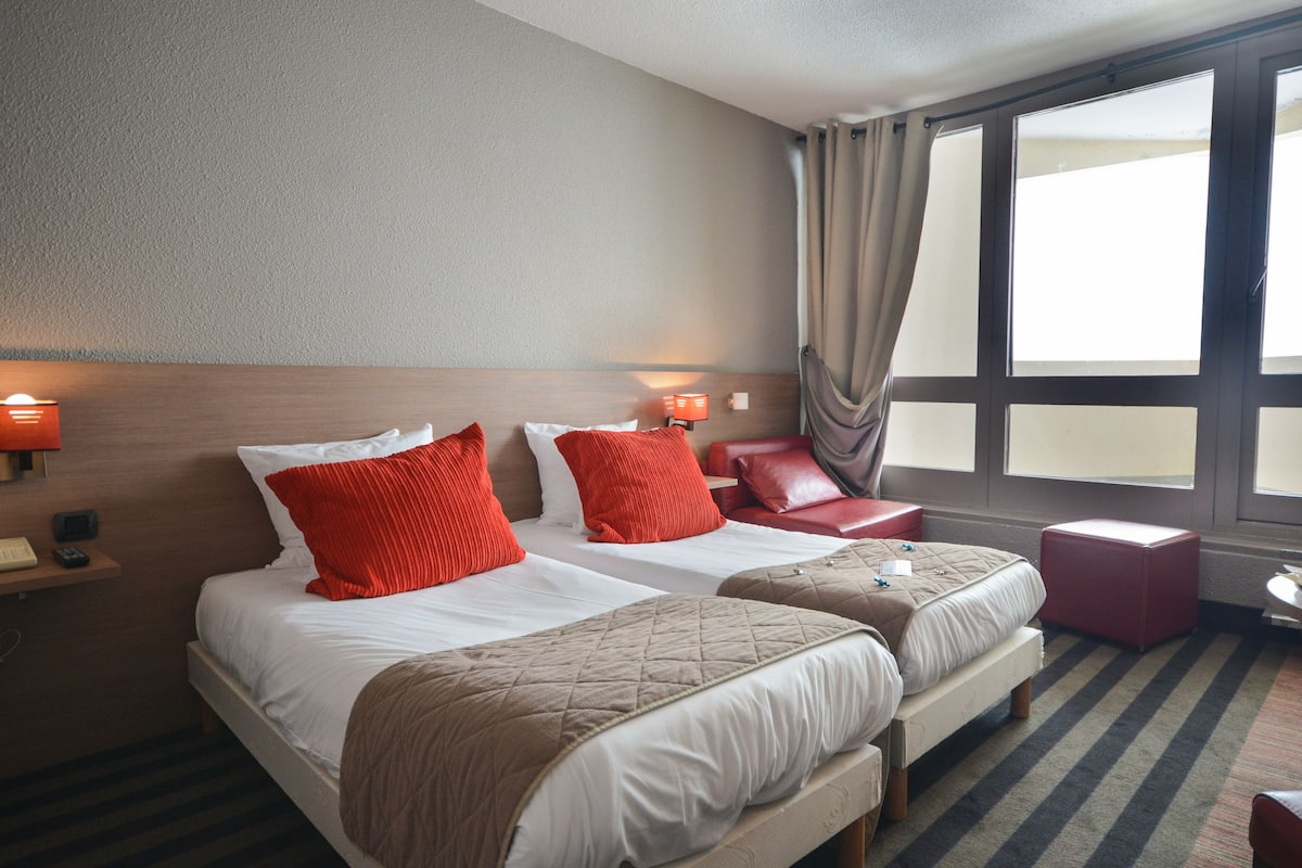 A hotel room with two beds, red pillows, a small table with breakfast, and a large window.