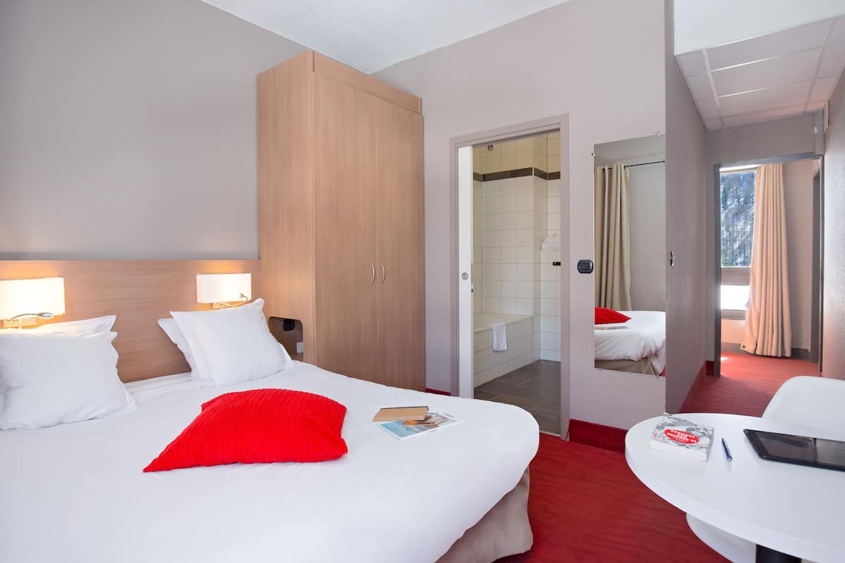 A hotel room with a bed, bedside lamps, a red pillow, a small table with a book and a pen, and a bathroom visible through an open door.