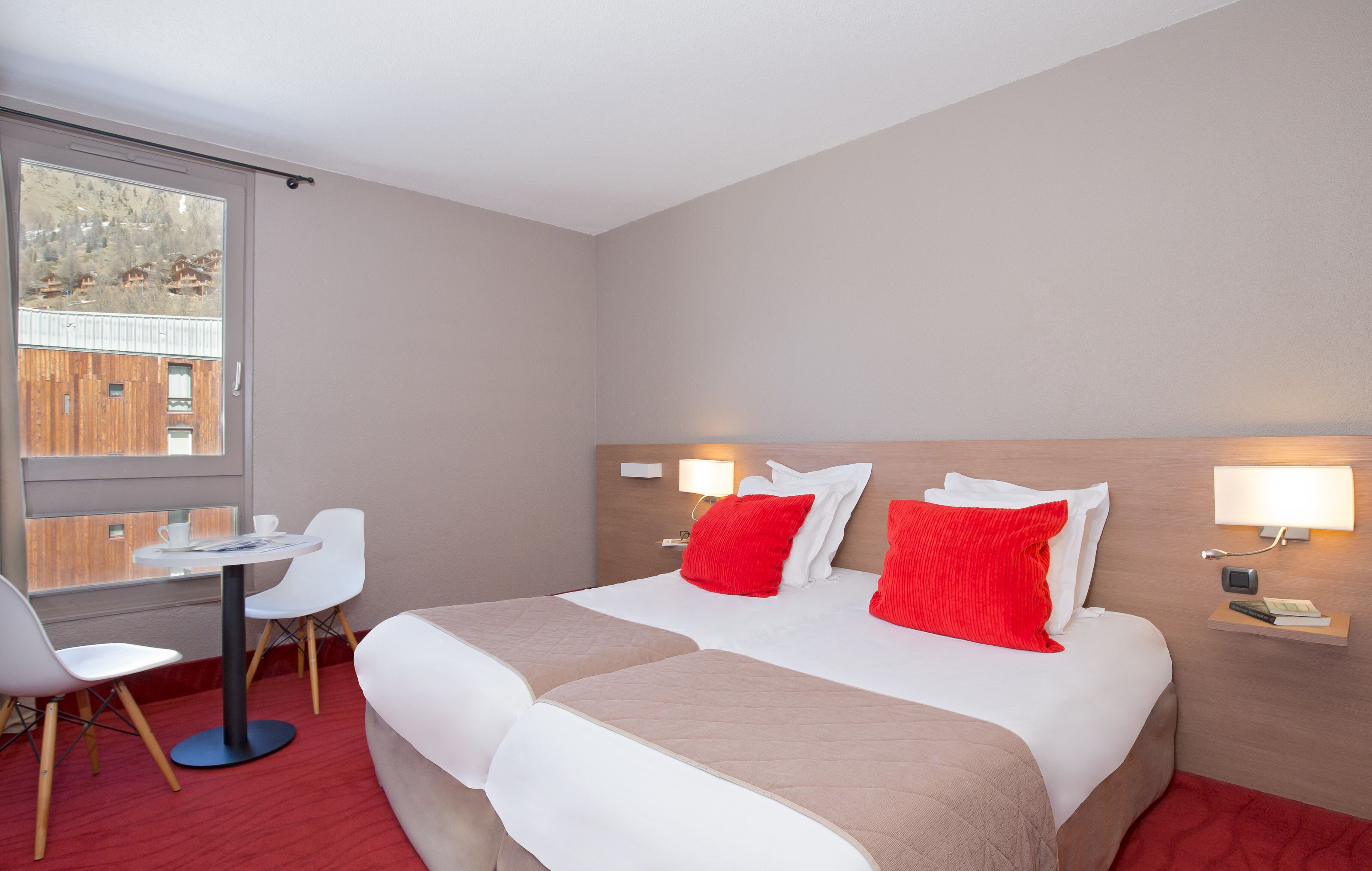 Habitación de hotel con cama, dos mesitas de noche con lámparas, un escritorio con silla y ventana con vistas a edificios.