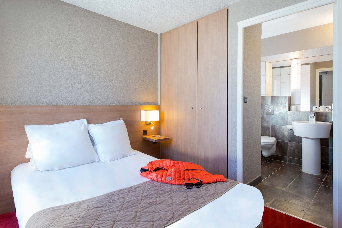 A hotel room with a bed, bedside lamps, a nightstand, a red jacket, and a bathroom with a sink and toilet.