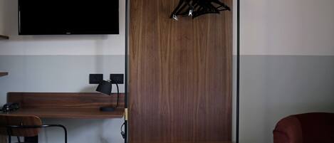 Une chambre d’hôtel avec un téléviseur fixé au mur, un bureau en bois avec une lampe, une chaise et un fauteuil rouge.