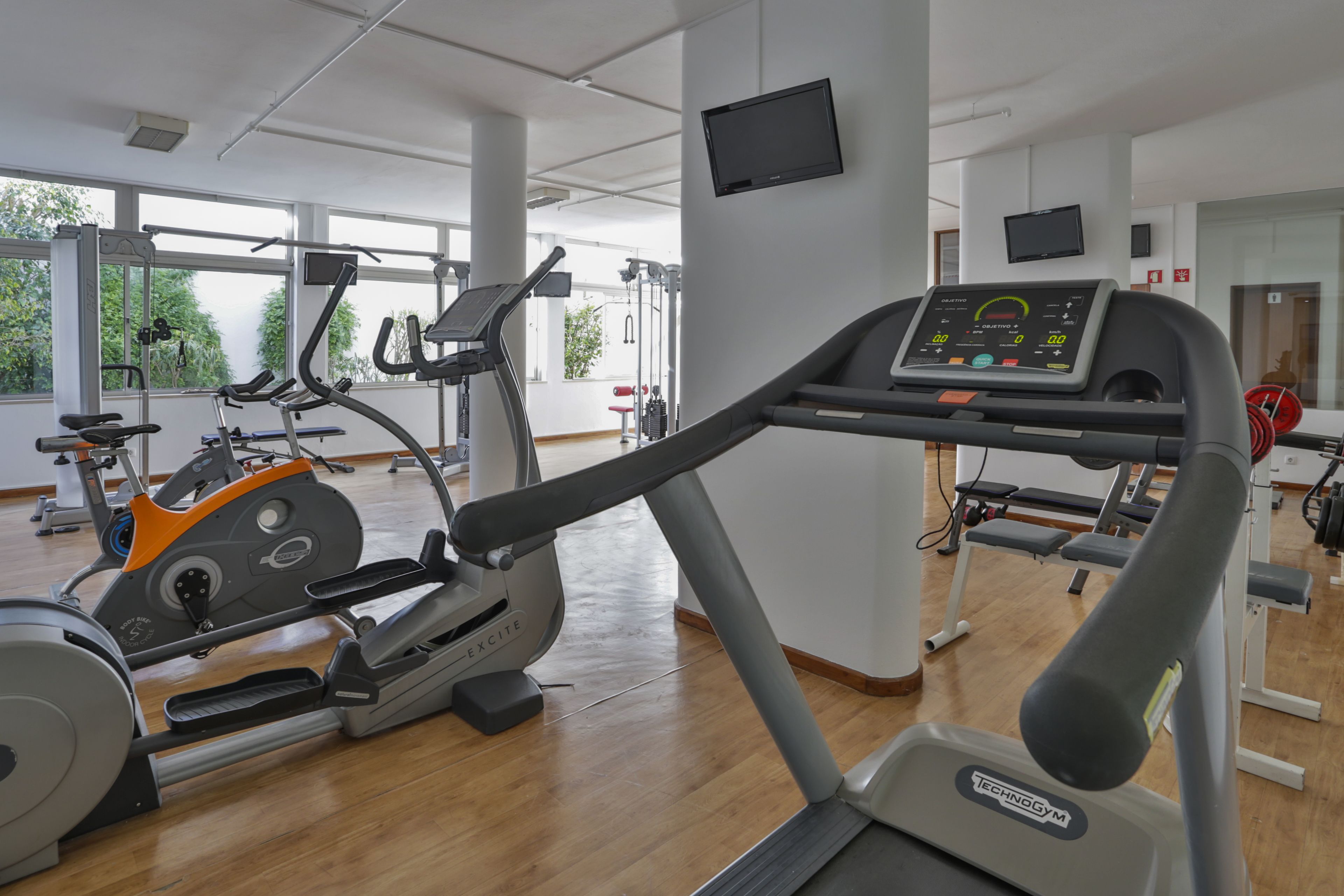 Un gimnasio con máquinas de cardio, un televisor y ventanas amplias.