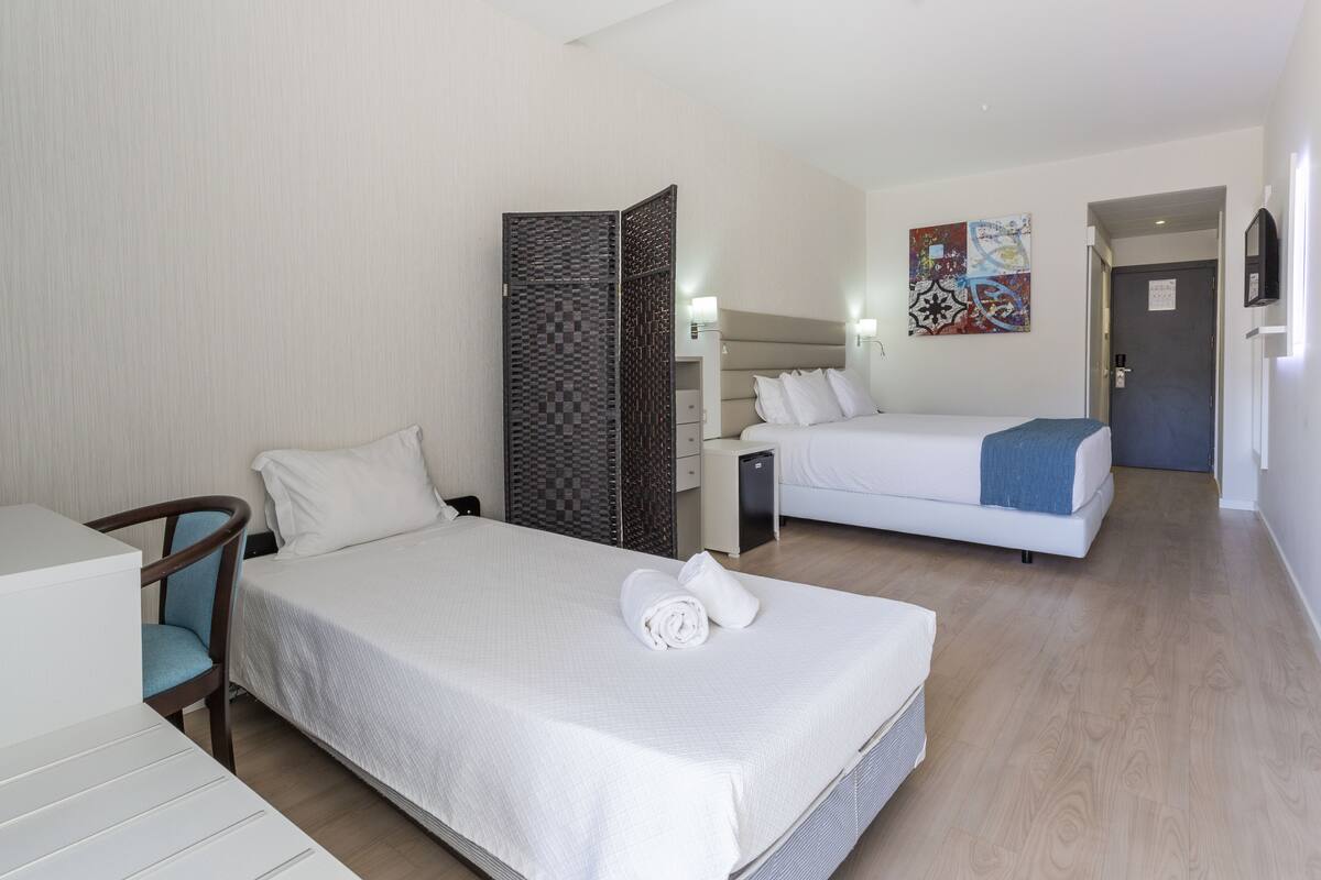 A hotel room with a large bed, a desk, a chair, a wardrobe, and a bedside table.