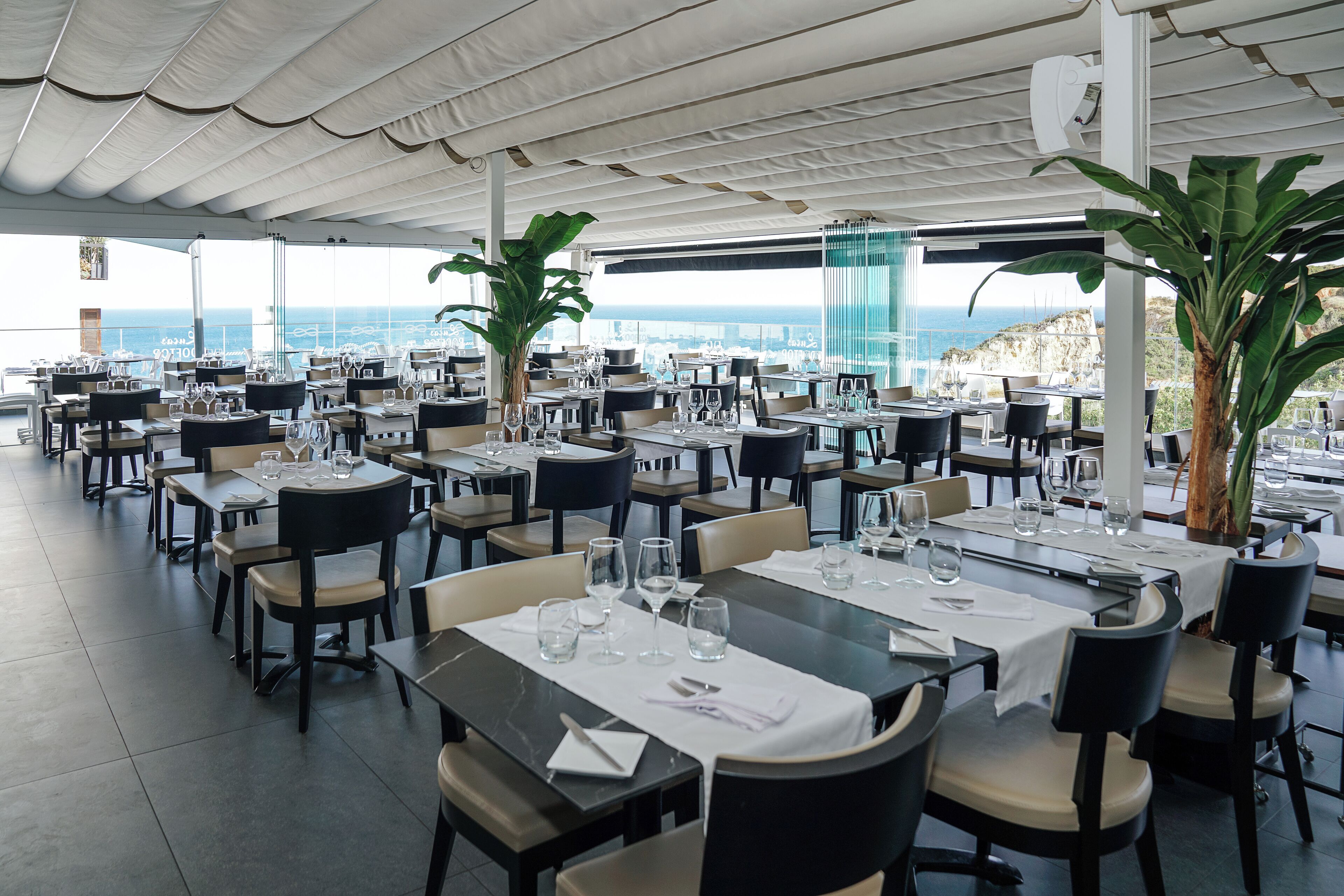 Un restaurant avec des tables dressées pour le service, de grandes plantes en pot et une vue sur la mer.