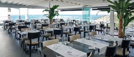Un restaurante con mesas dispuestas para comer, grandes macetas con plantas y vistas al mar.