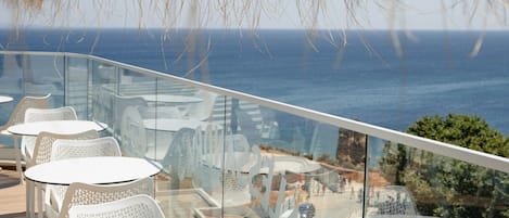 A glass-enclosed outdoor seating area with white chairs and tables, overlooking a coastal view.