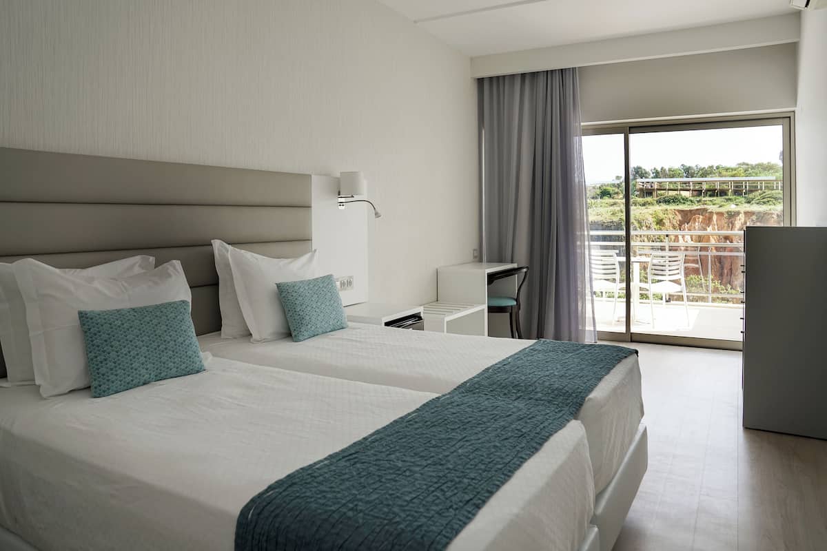 A hotel room with two beds, a desk, a chair, and a balcony with a view.